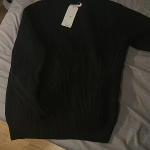 Svart ribbad tröja Maison Margiela - Svart ribbstickad tröja från Maison Margiela i storlek M. Tröjan har en klassisk rund halsringning och långa ärmar. Den är gjord i ett mjukt stickat material och har Margielas ikoniska vita stygn i nacken. Perfekt för dig som gillar minimalistisk och stilren design.