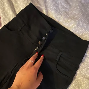 Svarta högmidjade jeans med knappar - Säljer ett par svarta högmidjade jeans med fyra silvriga knappar och dragkedja framtill. Byxorna har klassiska fickor fram och bak samt hällor för bälte. Snygg och enkel modell som passar till allt. Materialet känns som stretchig bomull eller jeans.
