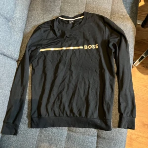 Svart HUGO BOSS tröja med guldtryck - Svart långärmad tröja från BOSS i bomull med stilrent guldtryck och logga framtill. Klassisk passform med rund halsringning och ribbade muddar. Perfekt för dig som gillar enkel men snygg design.