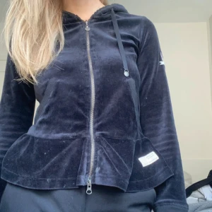 Svart sammetshoodie med dragkedja - Mysig svart hoodie från Odd Molly i mjuk sammet med dragkedja framtill, huva med snören och volangkant nertill. Liten broderad detalj på ärmen och en söt rosett bak. Perfekt för en chill och snygg look.