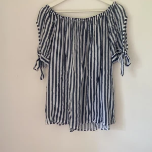 Randig offshoulder blus med knyt - Superfin blus med marinblå och vita vertikala ränder. Offshoulder-modell med resår upptill och korta ärmar som har knytband i sidorna. Luftigt tyg, perfekt för varma dagar och en chill look.