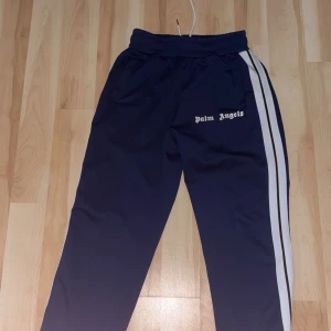 Palm Angels byxor S - Mörkblå byxor från Palm Angels med vita sidoränder och logga på vänster ben. Byxorna har resår i midjan med snörning och är gjorda i 100% polyester. Perfekta för en sportig och trendig look.