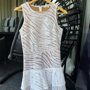 Vit ärmlös kortklänning med volang - Superfin vit kortklänning med vågigt broderat mönster och volang nertill. Klänningen är ärmlös och har ett innerfoder i beige! Perfekt för studenten! Köpt från Michael Kors med nypris på 4500kr men kom aldrig till användning, säljes därför. Passar XS/S.