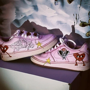 Nike Air Force 1 - Unika Nike Air Force 1 sneakers i ljusrosa med handmålade motiv som hjärtan, stjärnor och coola teckningar på sidorna. Skorna har vit sula, vita skosnören och klassisk rund tå. Perfekta för dig som vill sticka ut med färg och kreativ stil.