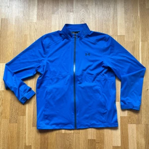 Under Armour Blå Regnjacka - Under Armour regnjacka i modellen Storm Rain Jacket och färgen blå. 20.000 mm vattenpelare! Storlek: L, Armhåla till Armhåla: 62 cm, Längd: 73 cm, Axel till Armöppning: 69 cm.  Material: 100% Polyester.