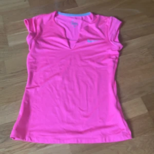 Rosa Dri-Fit träningstopp från Nike -  Rosa träningstopp från Nike med Dri-Fit-teknologi. Toppen har korta ärmar, v-ringning och en liten silverfärgad Nike-logga på bröstet. Storlek S. 
