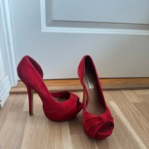 Röda mocka pumps från Steve Madden - Snygga röda pumps i mocka från Steve Madden med hög klack och öppen tå. Skorna har en elegant knutdetalj framtill och en klassisk siluett som verkligen sticker ut. 