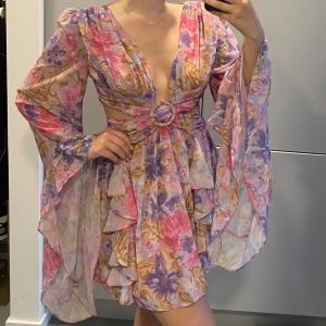 Blommig kortklänning med vida ärmar - Superfin kortklänning med blommigt mönster i rosa, lila och beige toner. Djup v-ringning, markerad midja med ringdetalj och cut-out, samt vida, fladdriga ärmar. Volangdetaljer och lätt, luftig känsla. Perfekt för sommarens alla tillfällen!