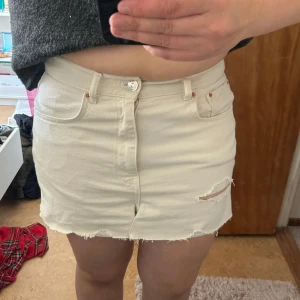 Vit jeans kjol med slitningar - Snygg vit kjol perfekt till sommaren. Kjolen har klassisk femficksdesign, slitningar framtill och en liten reva på ena benet för en trendig look. Perfekta för varma dagar och passar till allt.