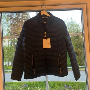 Moncler jacka  - Snygg svart dunjacka från Moncler med klassisk quiltad design och två dragkedjefickor framtill. Jackan har hög krage, Moncler-logga på ärmen och stilrena detaljer. Perfekt för dig som vill ha en clean och exklusiv look under kyliga dagar. Helt ny