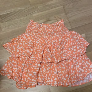Volangkjol från Samsa Collection - Superfin kort volangkjol från Samsa Collection i orange med vita blommiga mönster. Kjolen har bred smock i midjan och flera lager volanger som ger en riktigt söt och somrig vibe. Perfekt för dig som gillar färg och vill sticka ut lite extra!