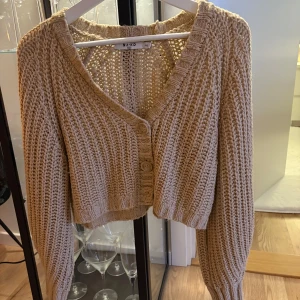 Beige stickad kofta från NA-KD - Säljer en snygg beige stickad kofta från NA-KD. Koftan är croppad med långa ärmar och har knappar framtill.