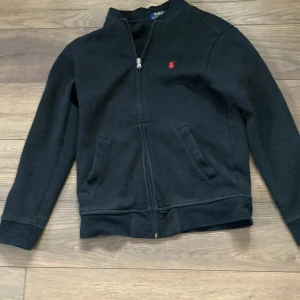 Svart tröja med dragkedja från Ralph Lauren - Säljer en stilren svart tröja från Ralph Lauren med dragkedja och det klassiska röda broderade logotypen på bröstet. Perfekt för en grischig och snygg look. Tröjan har långa ärmar och är tillverkad i ett mjukt material.