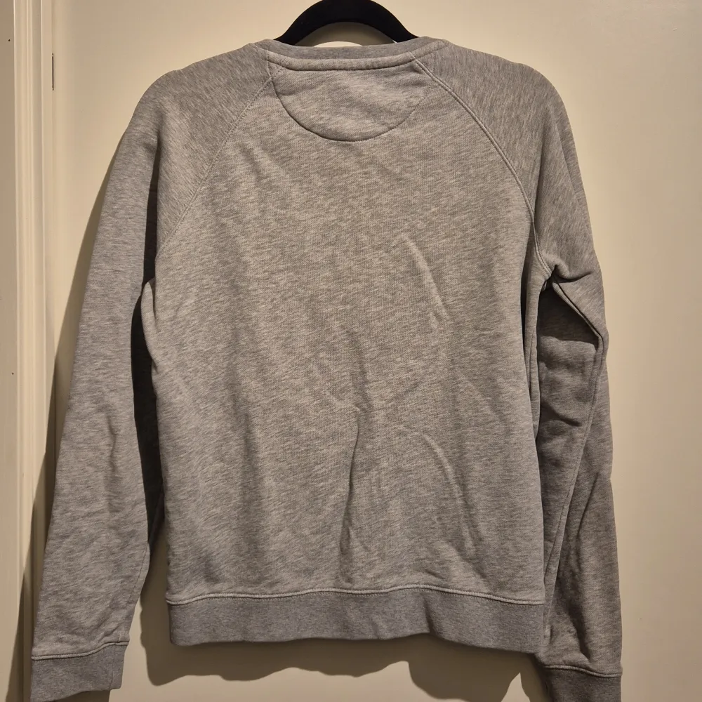 Säljer en stilren grå sweatshirt från GANT med broderad logga och ett emblem på bröstet. Tröjan har långa ärmar och rund halsringning. Perfekt för en avslappnad stil.. Neuletakit & Villapaidat.