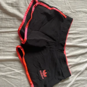 Svarta shorts från Adidas - Säljer ett par svarta shorts från Adidas med röd och rosa kant. De har en broderad Adidas-logga på framsidan och är perfekta för träning eller avslappnad stil. Shortsen är tillverkade i ett mjukt och bekvämt material.
