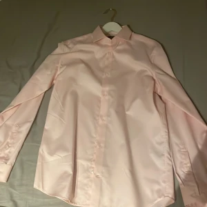 Ljusrosa skjorta - Elegant ljusrosa skjorta med klassisk krage och långa ärmar. Perfekt för en stilren look. Skjortan har knappar framtill och är tillverkad i ett mjukt material.