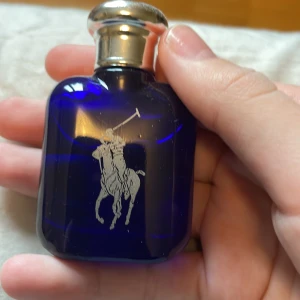 Blå parfymflaska från Ralph Lauren - Elegant blå parfymflaska från Ralph Lauren med en silverfärgad kork. Flaskan har en stilren design med en ingraverad polospelare på framsidan. Perfekt för den som söker en klassisk och tidlös doft.Mer än 14 ml kvar av 15