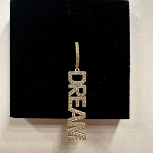 Guldörhänge med texten DREAM från Loaku - Helt oanvänt guldörhänge från Ioaku med texten 'DREAM' dekorerad med glittrande stenar. Säljer för att den inte kommer till användning då jag bara använder den andra. Den kom alltså i ett set med två örhängen men behövde bara ett så säljer den andra.