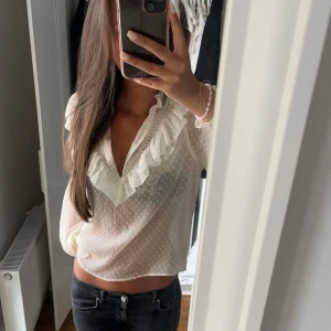 Vit blus med volangdetaljer - Säljer en söt vit blus med volangdetaljer och v-ringning. Blusen har ett diskret prickigt mönster och långa ärmar. Storlek m men passar mer xs-s