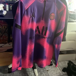 Lila och rosa tröja från Jordan x PSG - Säljer en cool lila och rosa tröja från Jordan i samarbete med PSG. Tröjan har en halv dragkedja och PSG-logga på bröstet. Perfekt för fotbollsfans som vill ha något unikt i garderoben.