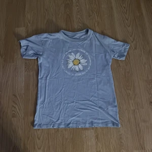 Blå t-shirt med blomtryck - Säljer en blå t-shirt med ett stort tryck av en blomma i mitten. T-shirten har korta ärmar och en rund halsringning. Perfekt för en avslappnad stil.