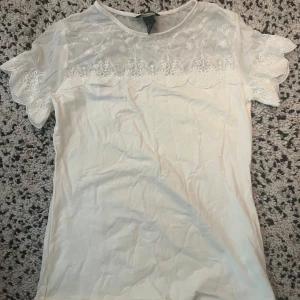 Vit blus från H&M - Säljer en söt vit blus från H&M med spetsdetaljer över bröstet och på de korta ärmarna. Perfekt för en somrig look eller en finare tillställning. Blusen har en rund halsringning och är gjord i ett lätt och bekvämt material.😍