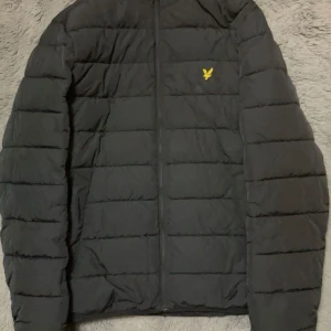 Svart pufferjacka från Lyle & Scott inte använd så mycket  - Säljer en svart pufferjacka från Lyle & Scott med dragkedja och deras ikoniska logga på bröstet. Jackan har långa ärmar och är perfekt för kyligare väder. Den är stilren och enkel att matcha med olika outfits.