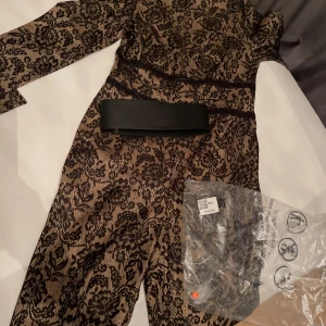 Svart jumpsuite med bälte  - Elegant svart och beige klänning med spetsmönster och långärmad design. Klänningen har en medföljande bälte för att framhäva midjan. Perfekt för en stilfull look. Helt ny aldrig använt, S.