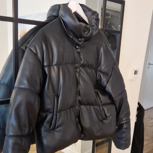 Svart pufferjacka - Snygg oversize svart pufferjacka med hög krage och knappar framtill. Jackan har en quiltad design och långa ärmar, perfekt för kyligare dagar. Den har en modern look och är både stilren och praktisk.