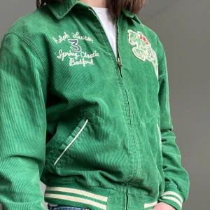 Grön manchesterjacka från Ralph Lauren - Säljer en grön manchesterjacka från Ralph Lauren med broderade detaljer och text på både fram- och baksidan. Jackan har dragkedja och ribbade muddar vid ärmslut och nederkant. Perfekt för en tidlös look!