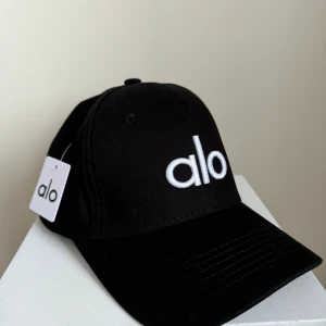 Svart keps från Alo - Snygg svart keps från Alo med vit broderad logotyp framtill. Kepsen har en klassisk design med justerbar passform och är perfekt för en stilren look.