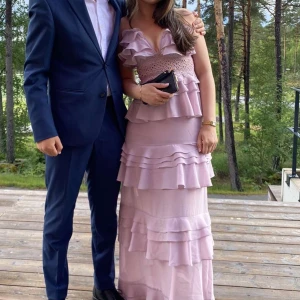 Ljuslila balklänning med volanger - Elegant ljuslila/rosa långklänning/balklänning med volanger och smala axelband. Klänningen har en vacker omlott-design upptill och flera lager av volanger. Perfekt för en bal. Endast använd en gång!! 