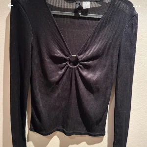 Svart ribbad topp med ringdetalj - Snygg svart ribbad topp med en unik ringdetalj framtill. Toppen har en djup v-ringning och långa ärmar, perfekt för en stilren look. Passar bra till både jeans och kjol.