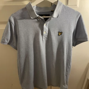 Ljusblå pikétröja från Lyle & Scott - Säljer en stilren ljusblå pikétröja från Lyle & Scott med klassisk krage och knappslå. Tröjan har en broderad logotyp på bröstet i gult och svart. Perfekt för en avslappnad men snygg look.