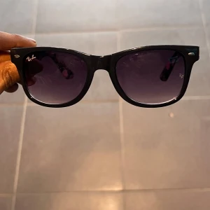  solglasögon med blommigt mönster från Ray-Ban - Snygga svarta solglasögon från Ray-Ban med ett coolt blommigt mönster på skalmarna. Perfekta för att ge en touch av stil till din look. Tillverkade i Italien med hög kvalitet.