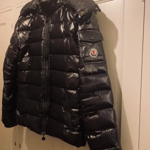 Svart dunjacka från Moncler - Snygg svart dunjacka från Moncler med glansig finish. Jackan har en löstagbar luva. Moncler-loggan är synlig på ärmen. 