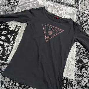 Snygg svart långärmad topp från Guess med det ikoniska triangellogotypen i rött på bröstet. Perfekt för en stilren och avslappnad look. Passar bra till jeans eller kjol.