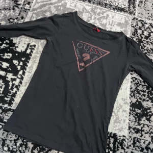 Svart långärmad topp från Guess - Snygg svart långärmad topp från Guess med det ikoniska triangellogotypen i rött på bröstet. Perfekt för en stilren och avslappnad look. Passar bra till jeans eller kjol.