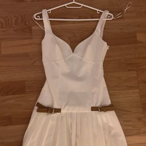 Vit klänning med bruna bälten från Zara - Snygg vit klänning med smala axelband och två bruna bälten i midjan. Klänningen har en dragkedja på sidan och ett plisserat kjolparti. Perfekt för sommarens alla tillfällen! Helt ny, aldrig använd då den är för liten.