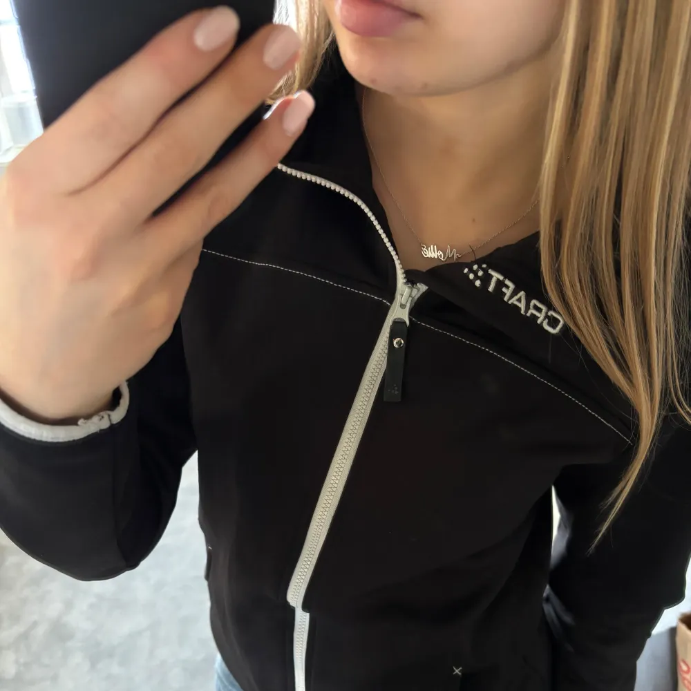 Snygg svart jacka från Craft med vit dragkedja och huva. Jackan har långa ärmar och praktiska fickor framtill. Perfekt för en sportig look eller en avslappnad dag ute.. Takit.