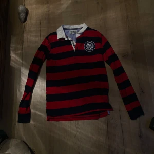 Randig pikétröja från Hampton Republic - Säljer en klassisk randig pikétröja från Hampton Republic i rött och svart med vit krage. Tröjan har långa ärmar och ett broderat emblem på bröstet. Perfekt för en stilren look.