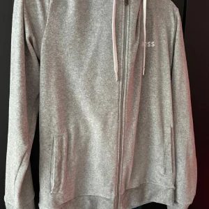 Grå huvtröja från Boss - Säljer en stilren grå huvtröja från Boss med dragkedja och justerbar huva. Tröjan har långa ärmar och praktiska fickor framtill. Perfekt för en avslappnad look.