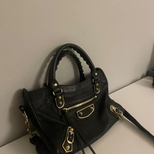 Svart handväska från Balenciaga - Snygg svart handväska från Balenciaga med guldfärgade detaljer och dragkedja. Väskan har en stilren design med både handtag och axelrem för flexibel användning. *Säljer den för den längre inte får någon användning, lågt pris för att äkthet inte kan garanteras då den är köpt secondhand.