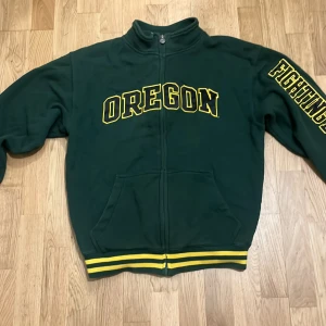 Grön hoodie med tryck - Säljer en grön hoodie med texten 'OREGON' och 'FIGHTING' i gult på framsidan. Baksidan har ett litet tryck med en maskot. Hoodien har dragkedja och ribbade muddar i gult och grönt.