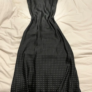 Svart prickig klänning - Snygg svart klänning med vita prickar och v-ringad design. Klänningen är ärmlös och har en elegant passform som framhäver figuren. Perfekt för en stilren look.