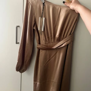 Beige satin klänning från NAKD - Elegant beige klänning i satin med en stilren oneshoulder-design. Klänningen har ett knytband i midjan för en smickrande passform och långa ärmar som ger en sofistikerad look. Perfekt för speciella tillfällen.