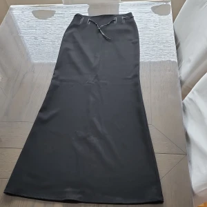 Svart långkjol från Maille Demoiselle - Elegant svart långkjol från Maille Demoiselle med en stilren design. Kjolen har en slits baktill och ett knytband i midjan för justerbar passform. Perfekt för en chic och sofistikerad look.