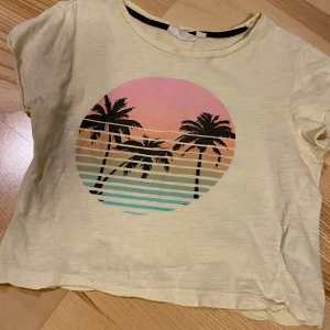 KappAhl gul t-shirt, storlek 134/140 - T-shirt med tryck av palmer mot en solnedgång i rosa, orange och blå toner. T-shirten har en gulaktig nyans och en rund halsringning med en svart kant.