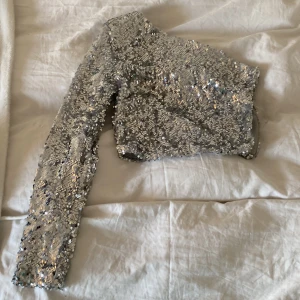 Silver paljettopp från Sinsay - Snygg silverfärgad paljettopp från Sinsay med en axel. Perfekt för en glittrig kvällslook. Toppen har en tight passform och är täckt med glittrande paljetter som fångar ljuset.