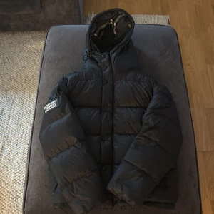 Svart pufferjacka från Burrybery - Säljer en svart pufferjacka från Burrybery med en cool logga på ärmen. Jackan har en quiltad design och en huva med ett rutigt mönster på insidan. Perfekt för kalla dagar med sin långa ärmar och dragkedja framtill. Hör av dig om du är intresserad! Man kan också göra den till en väst och 3/4 knappar är sönder